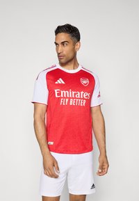 Rood en wit Adidas shirt met het logo "Emirates Fly Better", korte mouwen, ronde hals, een gladde textuur en een geometrisch patroon aan de voorkant.