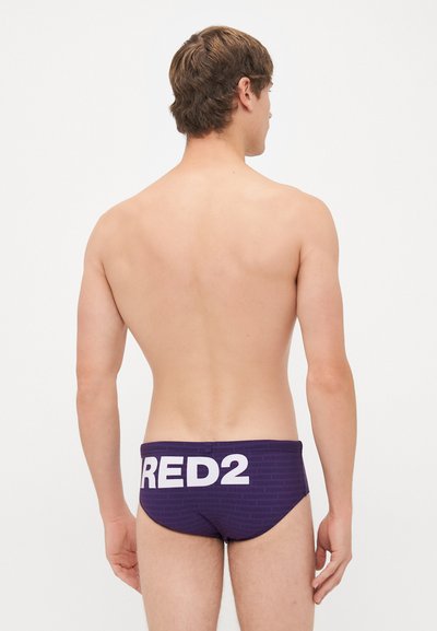Jeune homme de dos, portant un slip de bain violet foncé avec un grand texte blanc "RED2" sur la taille à l'arrière.