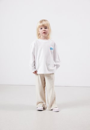 Junges Kind mit blonden Haaren, das ein weißes langärmliges Hemd, beigefarbene weite Hosen und weiße Turnschuhe trägt und vor einem weißen Hintergrund steht.