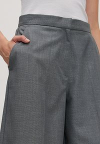 Persoon in een grijze, gestructureerde broek met een hand casual in de rechterzak, gecombineerd met een witte, geribbelde tanktop.