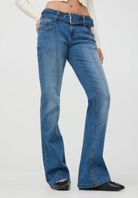 Nieco rozszerzane niebieskie dżinsy o średnim stanie, z paskiem i subtelnym detalem stębnowania wzdłuż bocznych szwów.