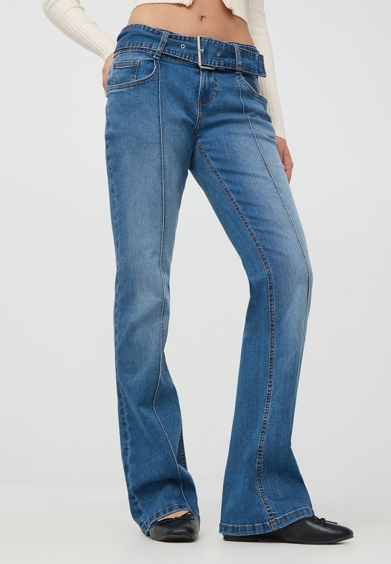 Nieco rozszerzane niebieskie dżinsy o średnim stanie, z paskiem i subtelnym detalem stębnowania wzdłuż bocznych szwów.