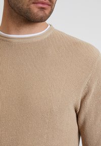 Pull en tricot beige avec un motif texturé, col rond et un détail de col intérieur blanc en contraste.