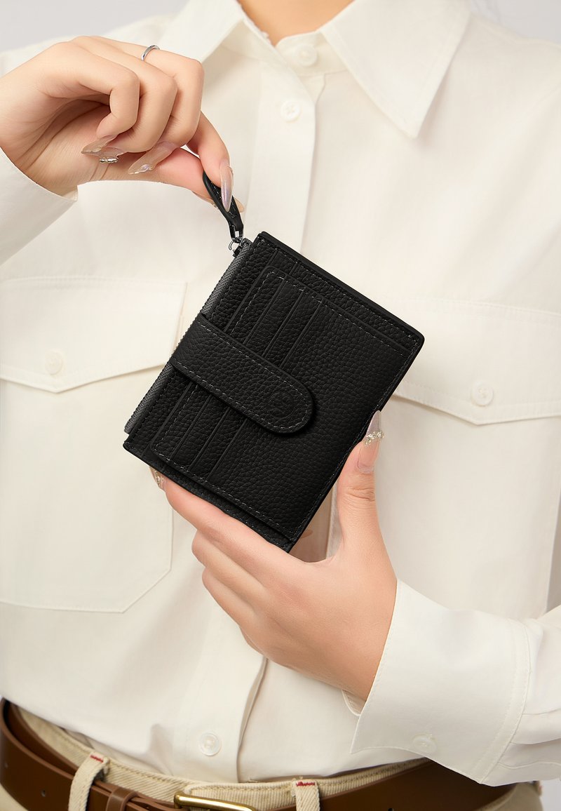 Portafoglio nero con texture, chiusura a scatto, slot per carte sul davanti e zip. Tenuto in mano contro una camicia di colore chiaro.