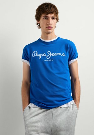 Giovane uomo che indossa una t-shirt blu Pepe Jeans London sopra una camicia bianca con pantaloni grigi, in piedi con le mani in tasca davanti a uno sfondo bianco.