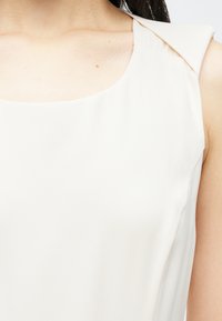 Ανοιχτό μπεζ αμάνικο τοπ με στρογγυλή λαιμόκοψη, που διαθέτει δομικές ενισχύσεις στους ώμους και ομαλό, υφασμάτινο υλικό.