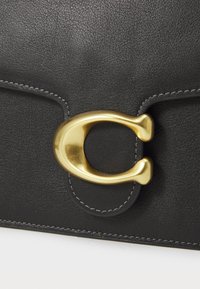 Sac en cuir noir avec un biais en métal doré en forme circulaire, avec des coutures visibles le long des bords. Texture lisse.