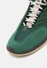 Green sneaker featuring a mix of mesh and suede materials, textured accents, black laces, and a tan rubber outsole.