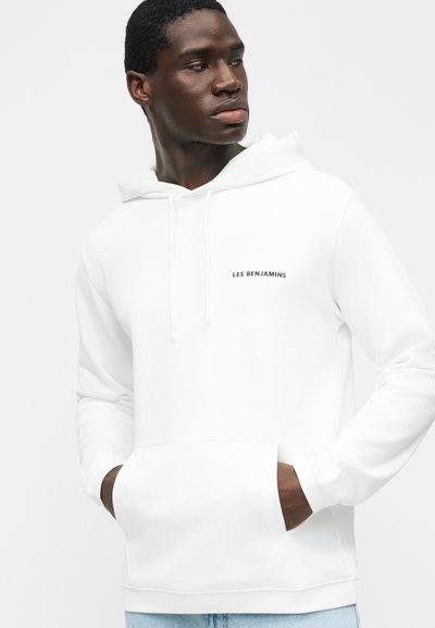 Hombre con sudadera blanca Les Benjamins con bolsillo delantero, mirando de lado contra un fondo claro y liso.