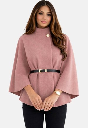 Femme aux longs cheveux portant une cape ceinturée rose poussiéreux avec des manches larges et un pantalon noir sur un fond uni.