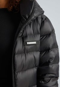 Giubbotto imbottito nero con design trapuntato, texture lucida, chiusura con zip e etichetta di marca sul petto. Aspetto minimalista e elegante.