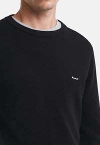 Schwarzer strukturierter Pullover mit einem gerippten Rundhalsausschnitt und einem kleinen weißen Logo. Über einem grauen Hemd getragen, das einen lässigen Schnitt hat.
