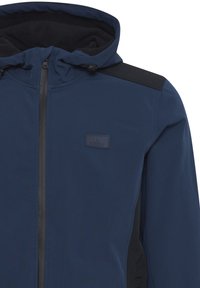 Zila zipa jaka ar kapuci, kurai ir melna pleca akcents, gluda tekstūra un mazs logotipa pārliktnis uz krūškurvja.