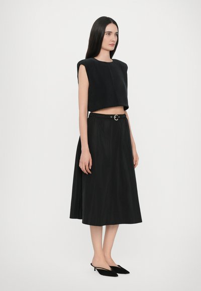 Zwarte cropped top met brede armsgaten, gecombineerd met een zwarte geplooide rok met een riem met een ronde gesp. Zwarte pumps met een spitse neus.