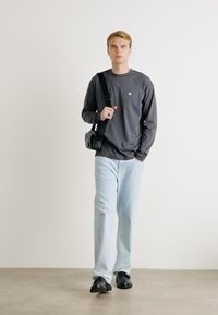 Šedá dlouhá košile, světle modré džíny, černé boty. Drží malou černou krosbody tašku. Jednoduchý design, hladká textilie.
