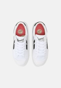 Hvite sneakers med svarte detaljer, snøring, teksturert stoff og rød innersåle. Har logo og sirkulært såleutseende.