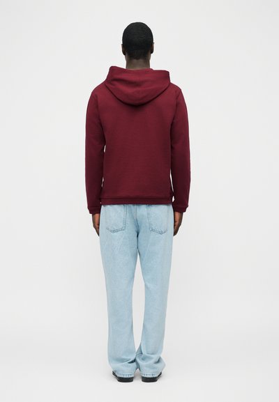 Άνδρας που φοράει μπορντό φούτερ με κουκούλα και ανοιχτόχρωμο τζιν, στέκεται γυρισμένος με την πλάτη σε έναν απλό ανοιχτό φόντο.