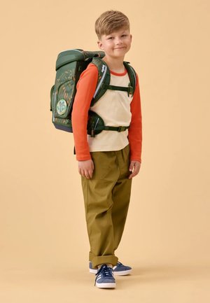 Grüner Rucksack mit gemusterten Akzenten, verstellbaren Trägern und einer Fronttasche. Wird von einem Kind in einem orange- und cremefarbenen Oberteil, grünen Hosen und blauen Schuhen getragen.