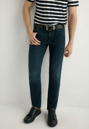 Mężczyzna ubrany w ciemnoniebieskie dżinsy, czarny pasek, czarne trampki oraz granatowo-białą pasiastą koszulę, stojący na jednolitym tle.