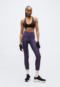 Femme en forme portant un soutien-gorge de sport noir, des leggings violets, des chaussettes blanches, des baskets noires et des gants sans doigts, debout devant un fond clair uni.