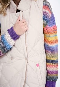 Beżowa kamizelka pikowana w diamentowy wzór, z kolorowo paskowanymi dzianinowymi rękawami w odcieniach fioletu, niebieskiego, różowego, zielonego i żółtego.