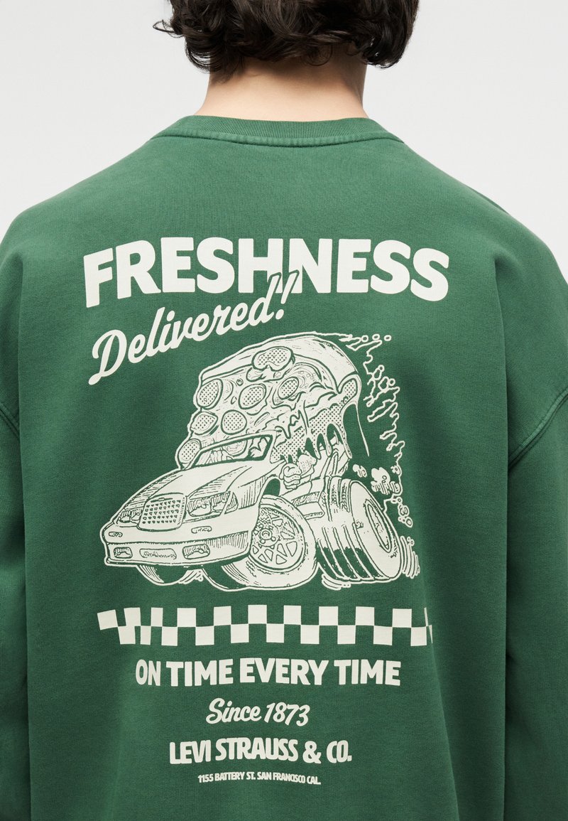 Sweatshirt vert avec un graphisme blanc représentant une voiture transportant une grande part de pizza, texte : « Fraîcheur livrée ! À l'heure à chaque fois, Levi Strauss & Co. »
