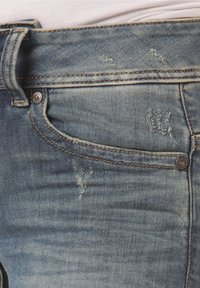Blå denimjeans med blekta områden och slitna detaljer runt framfickan och midjebandet.