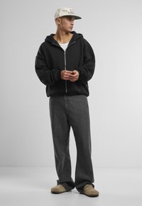 Schwarze Zip-Hoodie mit lockerer Passform, kombiniert mit grauen Jeans und hellbraunen Slippers. Das Modell trägt eine hellfarbige Mütze.