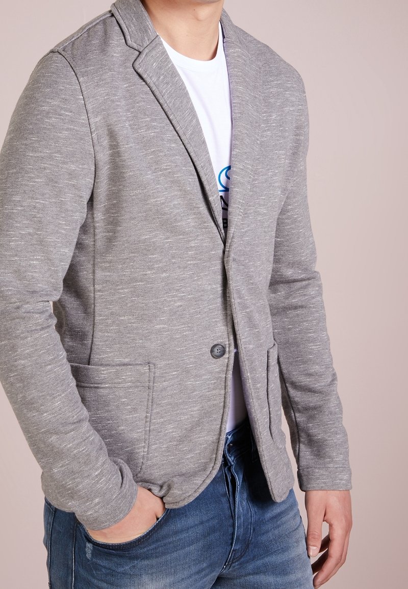 Mann trägt ein hellgraues, strukturiertes Sakko über einem weißen T-Shirt mit Grafik und blauer Jeans, steht mit einer Hand in der Tasche.