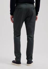 Mørkegrønne bukser lavet af tekstureret stof, med to baglommer med knapper. Båret med mørke slip-on sko.