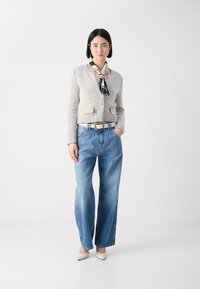 Lichtgrijze herringbone cardigan met knopen, blauwe wijde jeans en een zwart-wit sjaal die over de hals is gedrapeerd. Witte hakken.
