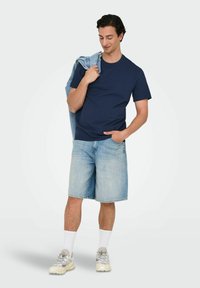 Mørkeblå t-shirt, lyseblå denimshorts og grå sneakers. En denimjakke hænger over den ene skulder. Hvide sokker er synlige.