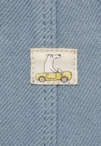 Label met een cartoonbeer in een gele auto op een crèmekleurig stuk, genaaid op lichtblauwe textuurstof met een visgraatpatroon.