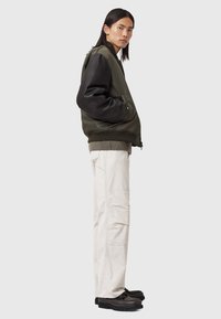 Jeune personne aux longs cheveux noirs portant une veste bomber verte et noire, un pantalon cargo beige clair, et des chaussures marron foncé, se tenant de côté.