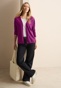 Gilet fuchsia sur un t-shirt blanc, associé à un jean en denim foncé et des baskets blanches. Le modèle tient un sac fourre-tout en crème matelassé.