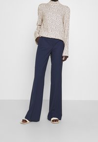 Donna che indossa pantaloni a gamba larga blu navy, una blusa a maniche lunghe beige con pois e sandali aperti beige, in piedi contro uno sfondo bianco.