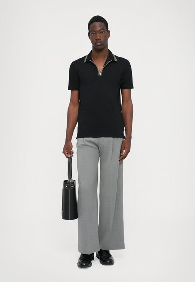 Camisa polo negra con cuello con cremallera, pantalones anchos gris, y zapatos negros. Sosteniendo una bolsa cilíndrica negra con una correa.