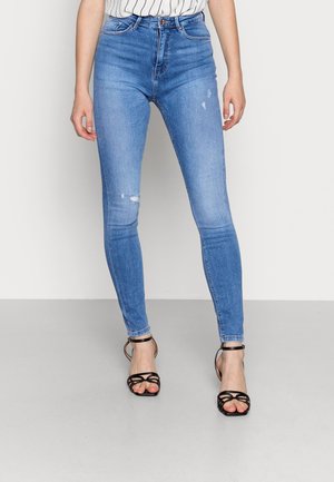 Slim fit blauwe jeans met lichte slijtage, gedragen met zwarte sandalen met hoge hakken en bandjes, op een persoon die staat tegen een effen achtergrond.