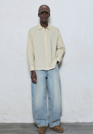 Jeune homme portant une chemise beige rayée à boutons, un jean large bleu clair, une casquette beige et des chaussures marron, debout contre un mur clair.