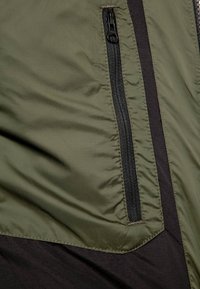 Giacca leggera verde oliva con una tasca con cerniera nera sul lato sinistro; presenta una texture liscia e un pannello scuro a contrasto sottostante.