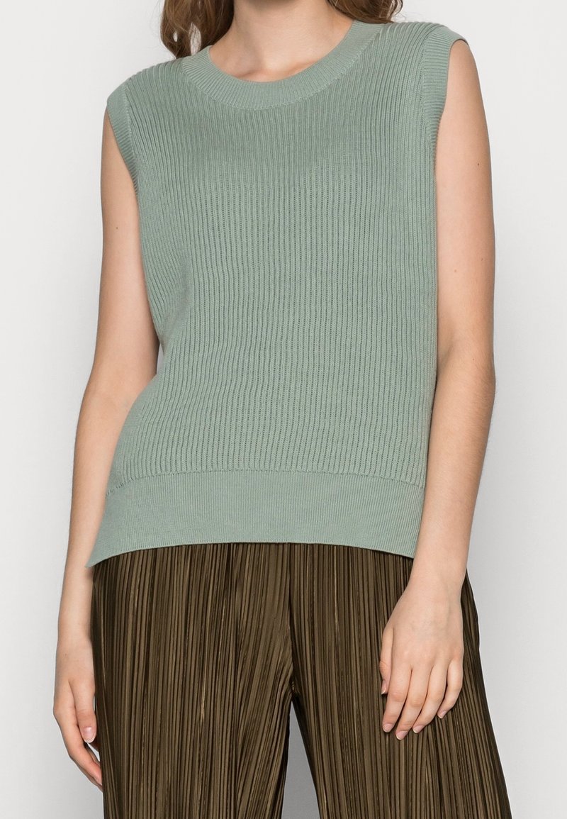 Femme portant un haut sans manches en maille côtelée vert clair associé à un pantalon plissé marron taille haute.