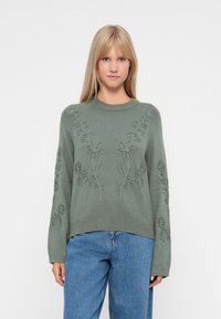 Pull en maille verte avec broderie florale, col rond et manches longues, associé à un pantalon en denim bleu. Texture décontractée et coupe ample.