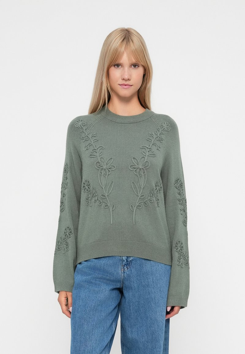 Pull en maille verte avec broderie florale, col rond et manches longues, associé à un pantalon en denim bleu. Texture décontractée et coupe ample.