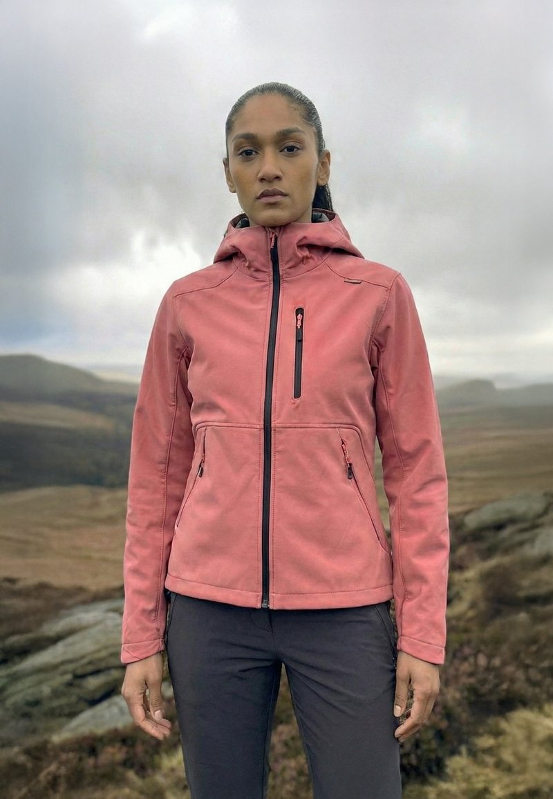 Femme debout en plein air sur un paysage rocheux et herbeux, portant une veste rose zippée et un pantalon noir sous un ciel couvert.
