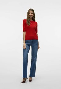 Röd kortärmad tröja med ribbad textur, i kombination med blå utsvängda jeans och mörkröda högklackade skor. Enkel bakgrund.