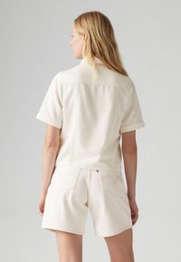 Kremfarget cropped skjorte med korte ermer, med en glatt tekstur og en enkel krage. Kombinert med matchende avkuttede shorts.