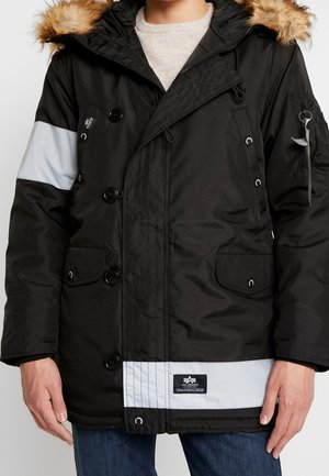 Czarna zimowa parka z kapturem obszytym futrem, zapięciem na guziki i suwak, odblaskowymi paskami na rękawie i przy dolnym brzegu, noszona na jasnym swetrze i dżinsach.