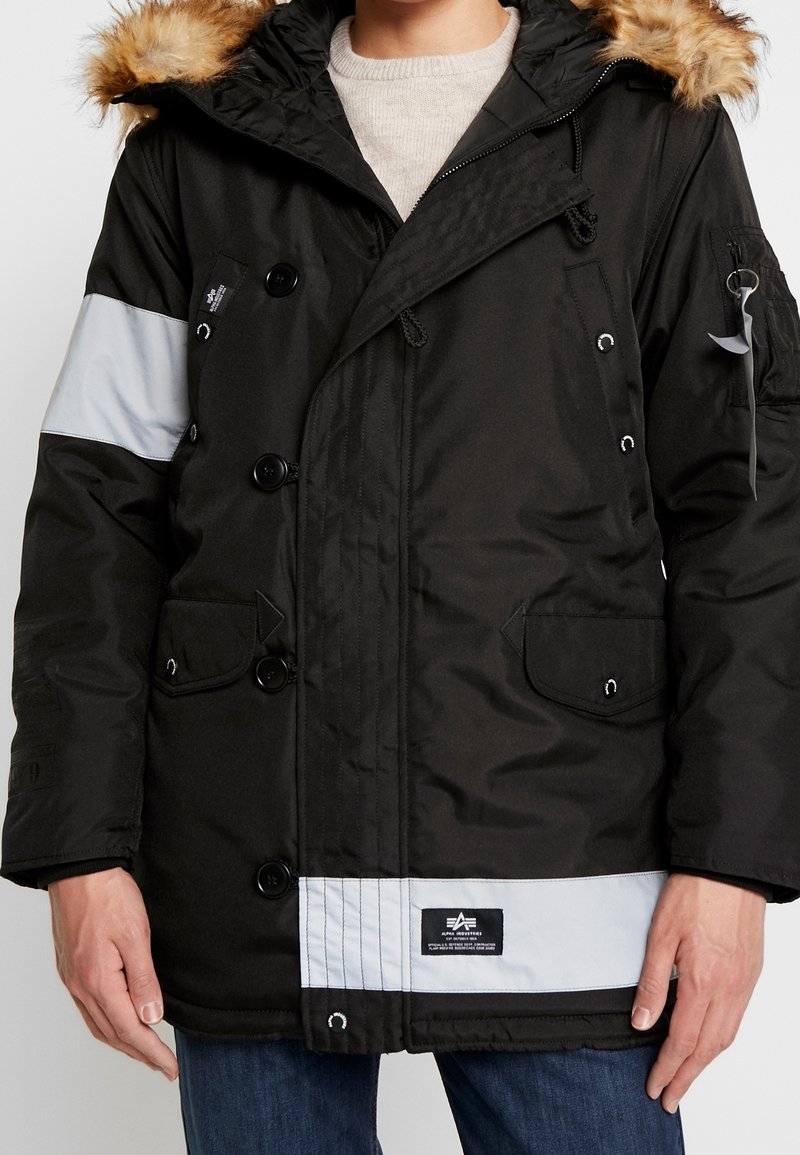 Parka d'hiver noire avec capuche bordée de fourrure, fermeture par boutons et fermeture éclair, bandes réfléchissantes sur la manche et l'ourlet, portée par-dessus un pull léger et un jean.