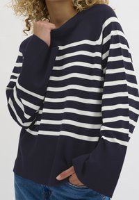 Modell i en marinblå och vit randig tröja med vida ärmar. Hon håller sin vänstra arm nära bröstet, mot en enkel bakgrund.