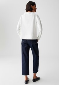 Vrouw in een textuurloze witte top met lange mouwen, donkerblauwe cropped wijde jeans en zwarte platte schoenen, staande tegen een effen achtergrond.